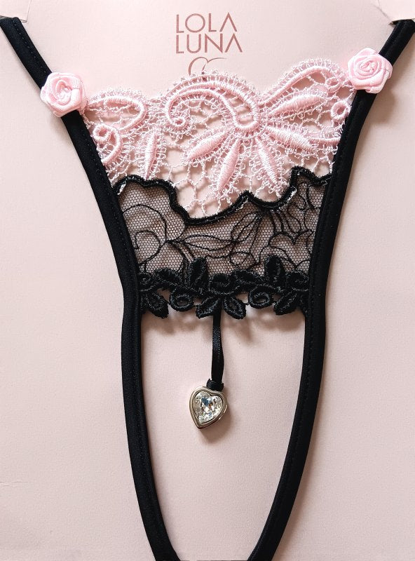 Mini string noir ouvert à l’entrejambe, orné de dentelle noire et rose délicate, avec un bijou en forme de cœur placé au centre.