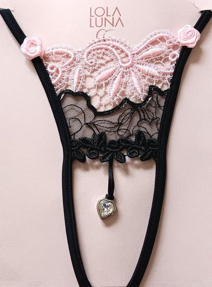 Mini string noir ouvert à l’entrejambe, orné de dentelle noire et rose délicate, avec un bijou en forme de cœur placé au centre.